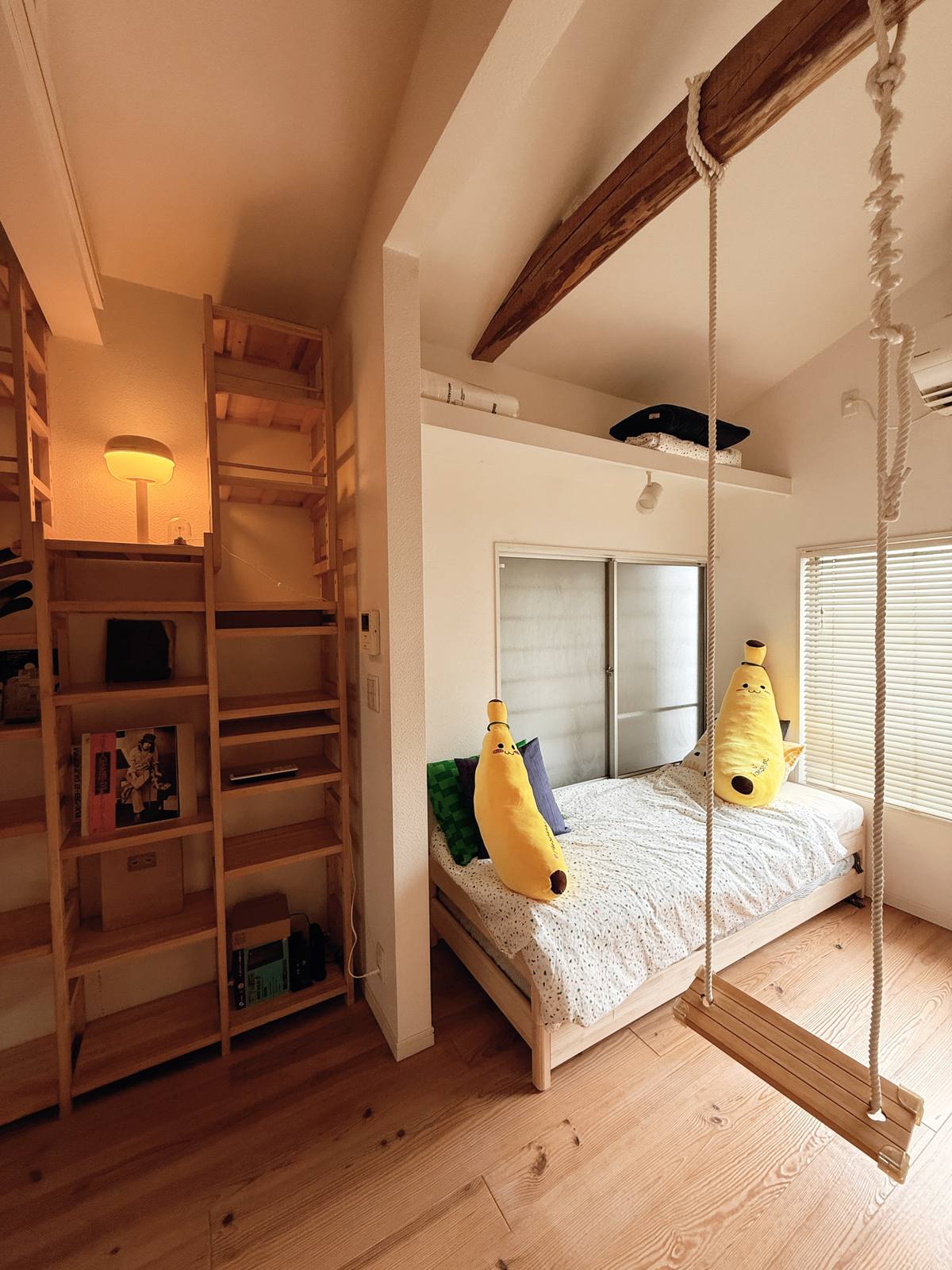 A cosy bedroom nook with a wooden rope swing, banana plushies on the bed and a book-lined shelving ladder, in warm afternoon light.